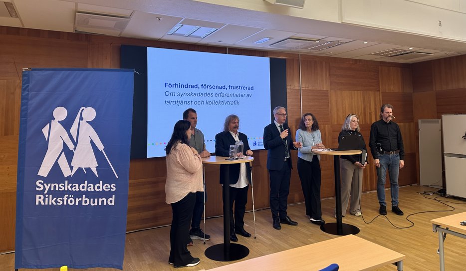 Sju personer står i en sal framför en stor skärm med texten "Förhindrad, försenad, frustrerad - Om synskadades erfarenheter av färdtjänst och kollektivtrafik".