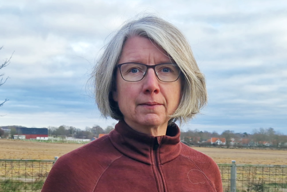 En kvinna står framför en äng i ett höstigt landskap. Hon har kort grått hår klippt i page, en röd funktionströja och gladögon med svarta bågar. Kvinnan ser med allvarlig blick in i kameran.