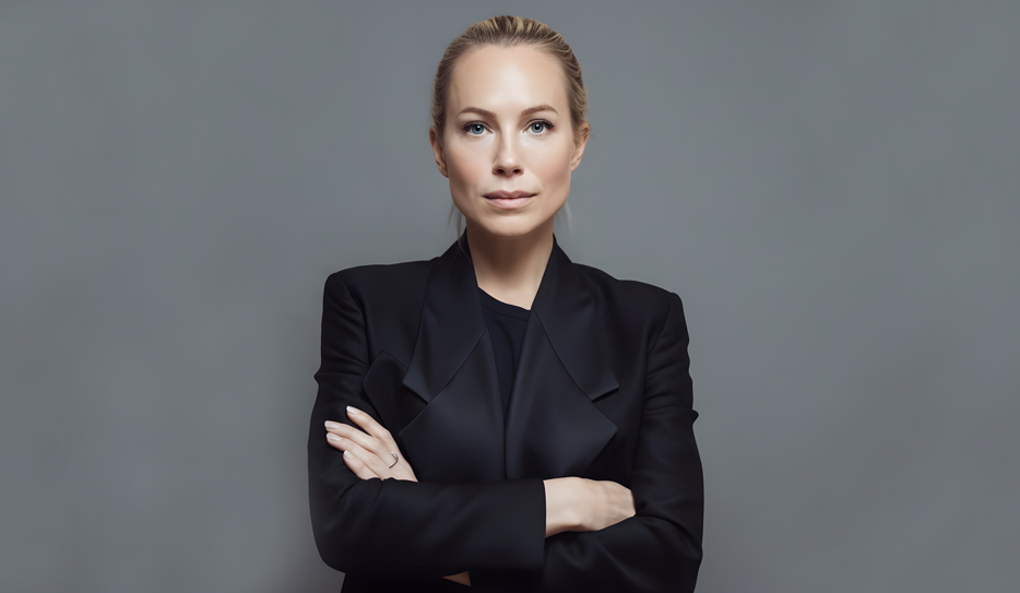 Anna Quarnström står med armarna i kors och tittar rakt in i kameran. Hon är klädd i svart kavaj och har sitt blonda hår uppsatt i en knut i nacken.