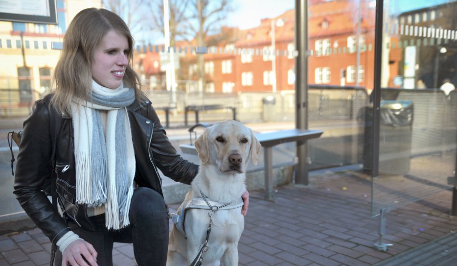 Viktoria sitter på knä bredvid sin ledarhund Poppe som är en ljus jaktlabrador. Viktora är i 25-årsålden och har skinnjacka och halsduk, hon har blicken riktad mot Poppe och ser glad ut.