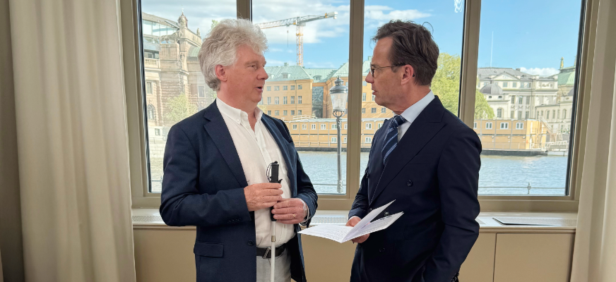 SRF:s förbundsordförande Niklas Mattsson och Ulf Kristersson (M) står framför ett stort fönster i Rosenbad med utsikt över Riksdagen och vattnet. Båda är klädda i mörka kostymer och samtalar engagerat. I Kristerssons hans syns SRF:s valbroschyr och Niklas Mattson håller i sin vita käpp.