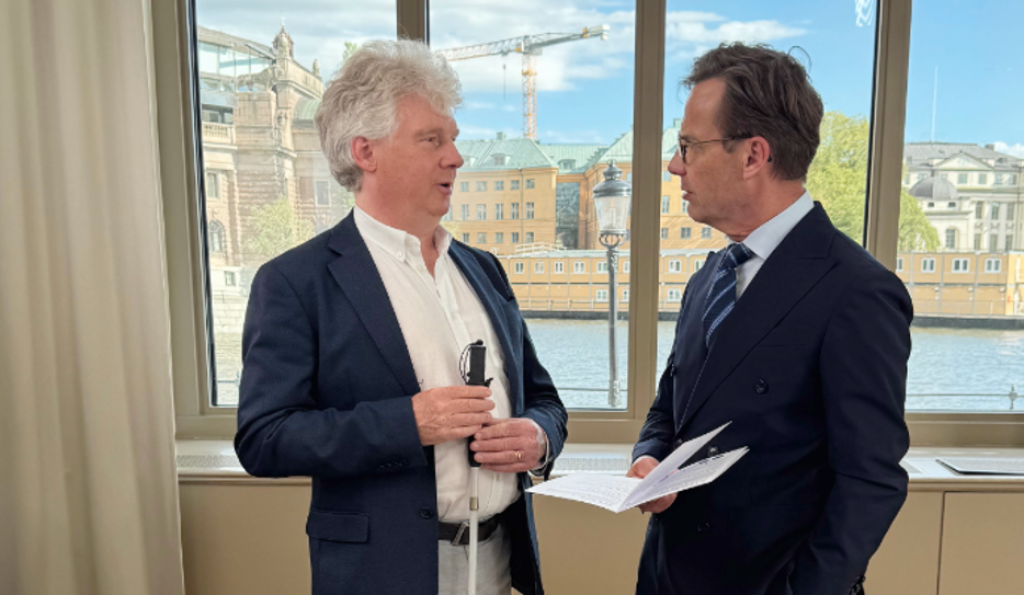 SRF:s förbundsordförande Niklas Mattsson och Ulf Kristersson (M) står framför ett stort fönster i Rosenbad med utsikt över Riksdagen och vattnet. Båda är klädda i mörka kostymer och samtalar engagerat. I Kristerssons hans syns SRF:s valbroschyr och Niklas Mattson håller i sin vita käpp.