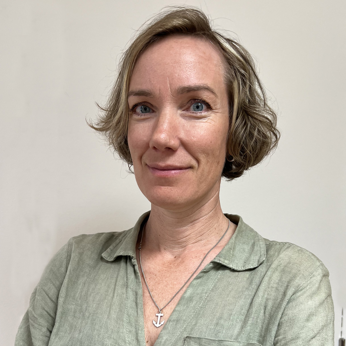 Leende kvinna i 50-årsåldern, rågblond page med självfall, ljusgrön linneskjorta, halsband med ankare som berlock.
