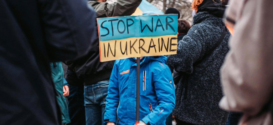 En folksamling deltar i en demonstration utomhus. I förgrunden står ett barn i blå jacka och håller en skylt med texten ”STOP WAR IN UKRAINE”. Bakom barnet syns människor med vinterkläder.