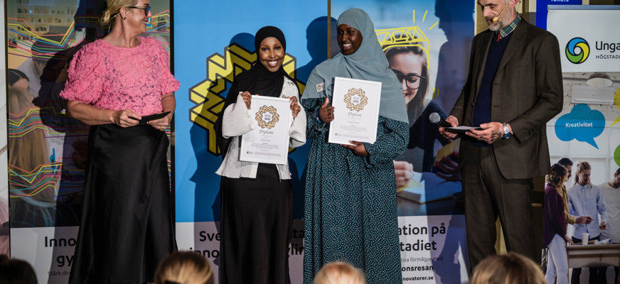 Hodan Yusuf och Deeka Sheikh står på scen med varsitt diplom i handen. De ler stort och i bakgrunden syns affischer med texten ”Unga innovatörer”.