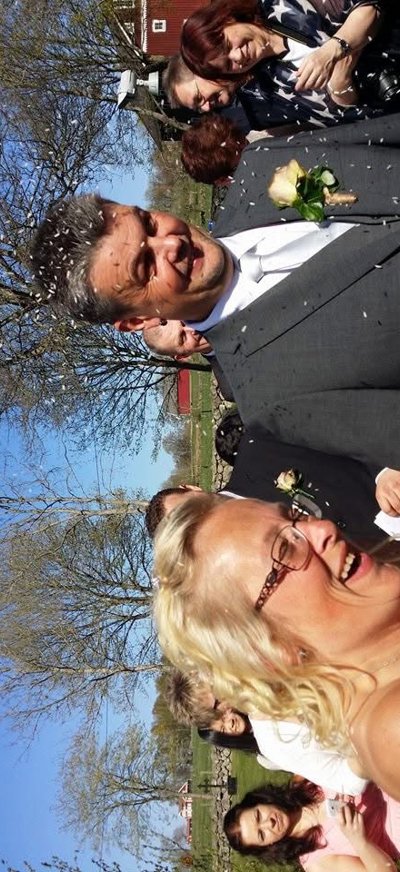 Ett nygift par går utomhus medan ris kastas över dem. De skrattar, och bruden håller en bukett. Flera gäster syns i bakgrunden under klarblå himmel.