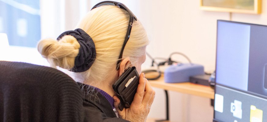 En kvinna med blont hår uppsatt i en knut sitter och pratar i en mobiltelefon. Hon har solglasögon uppe på huvudet och hörlurar runt halsen. Fotot är en genrebild.