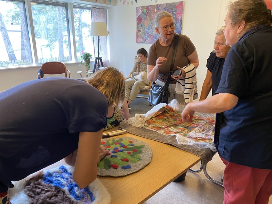 Bilden visar en grupp människor som deltar i ett ulltovningsprojekt i en gemensam eller workshopmiljö. Personerna arbetar med färgglada ullstycken utlagda på ett bord, för att skapa tovat material. Rummet har stora fönster som släpper in naturligt ljus och är dekorerat med konstverk och vimplar. Det är en kreativ och social aktivitet i en ljus och inspirerande miljö, där deltagarna engagerar sig i hantverket ulltovning.