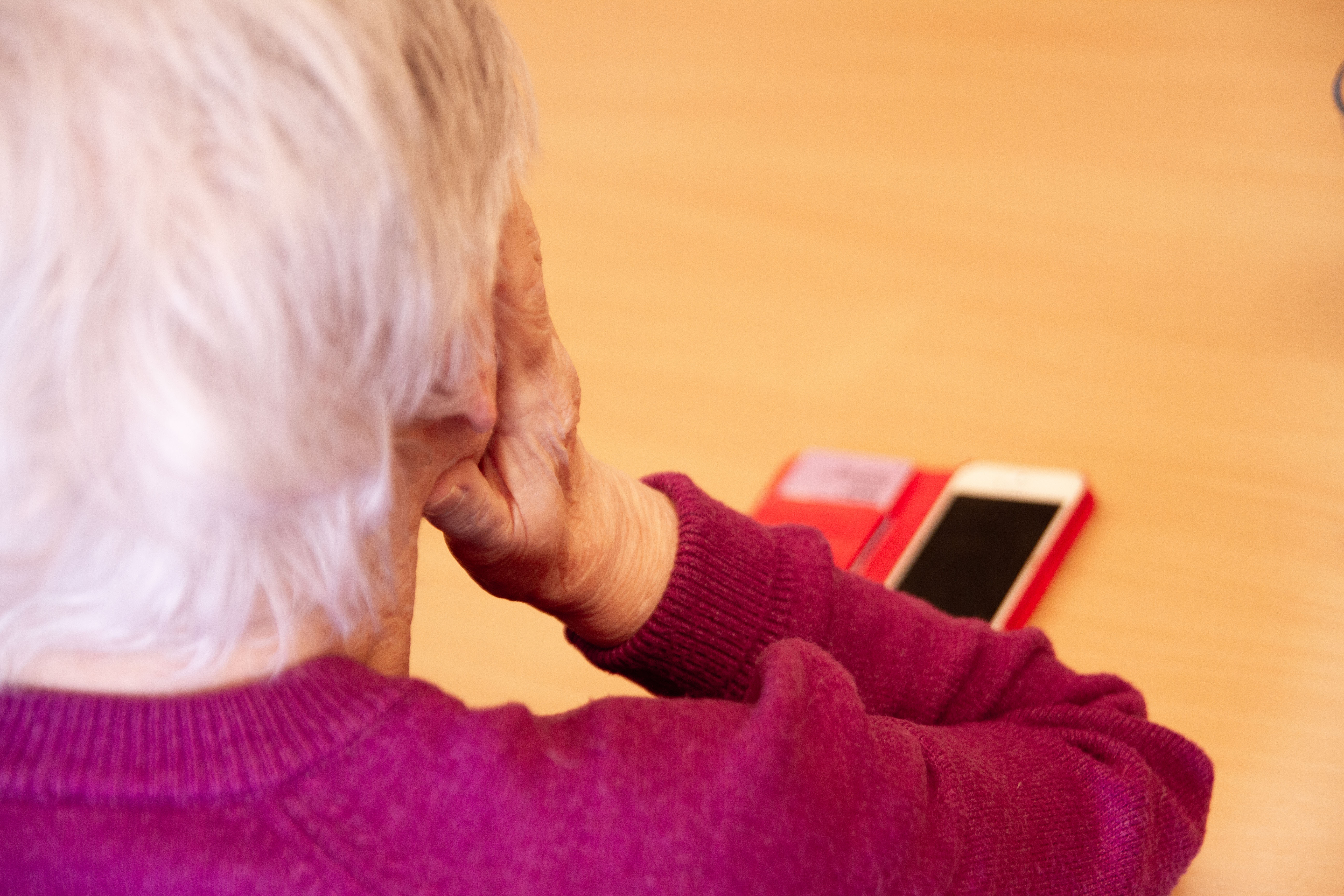 Äldre dam sedd snett bakifrån. Hon lutar huvudet i händerna. Framför henne på bordet syns en mobiltelefon.