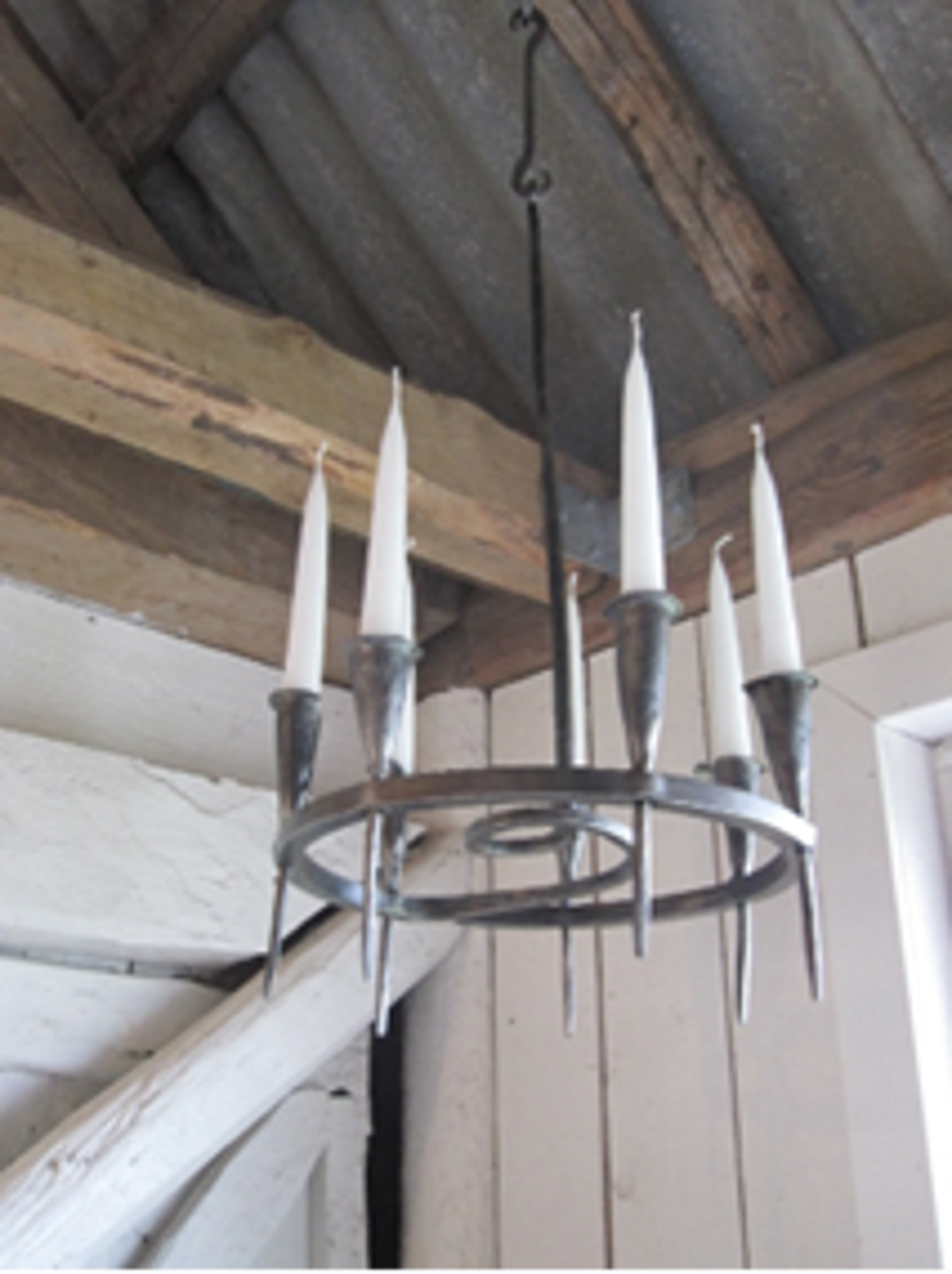 Ljuskronan är en rund, metallkonstruktion med tydlig rustik karaktär. Den hänger från taket med hjälp av en stadig metallstång och krok, vilket ger ett robust och industriellt uttryck. Ringen bär sex höga, vita stearinljus, jämnt fördelade runt cirkeln, vilket förstärker dess symmetriska och balanserade form. Designen är enkel men effektfull – utan elektriska ljuskällor, vilket ger en gammaldags charm. Kombinationen av metall och levande ljus skapar en varm atmosfär som passar perfekt i lantliga eller historiska miljöer. Ljuskronan är både funktionell och dekorativ, och dess minimalistiska stil gör den till ett blickfång i rummet.