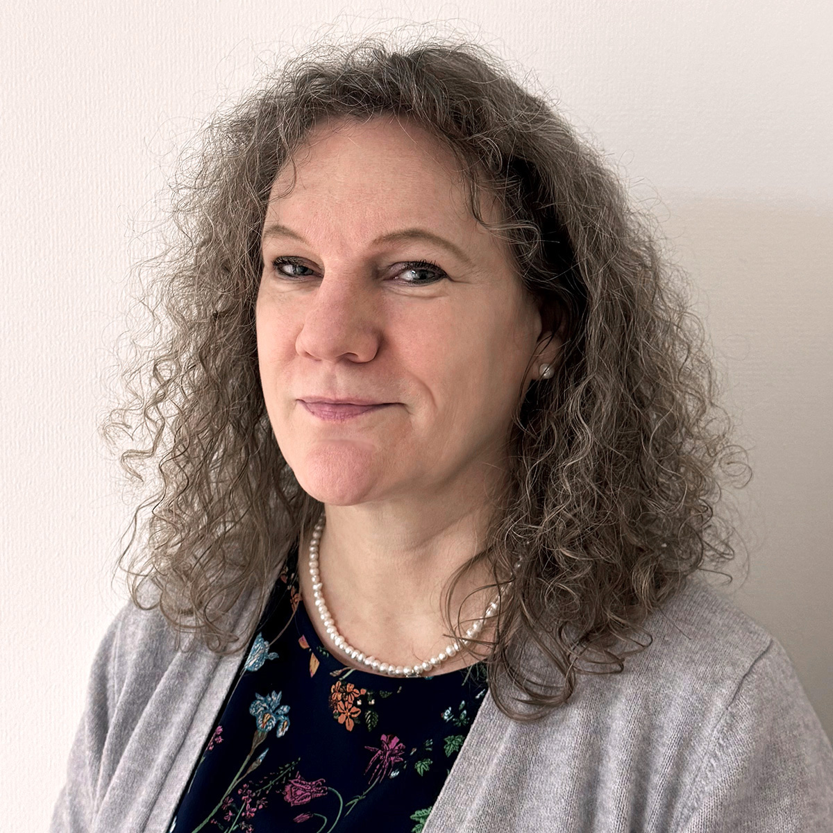 Portättbild av Jessica Helin. Jessica har axellångt hår som är lockigt och mörkt med grå slingor. Hon är klädd i en svart tröja med blommor och en grå kofta. Runt halsen och i öronen har hon vita pärlor.