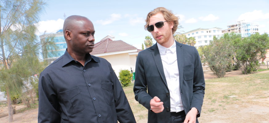 Två män samtalar utomhus. Ena mannen är svart och har rakat huvud. Han är klädd i svart skjorta och svarta byxor och håller en hopvikt vit käpp i handen. Den andra mannen är vit och har rödlätt hår och skägg. Han är klädd i mörk kostym med vit skjorta under och bär mörka solglasögon. De befinner sig i ett Afrikanskt land. Marken är torr och sandig och runt dem syns vita hus och gröna träd och buskar.