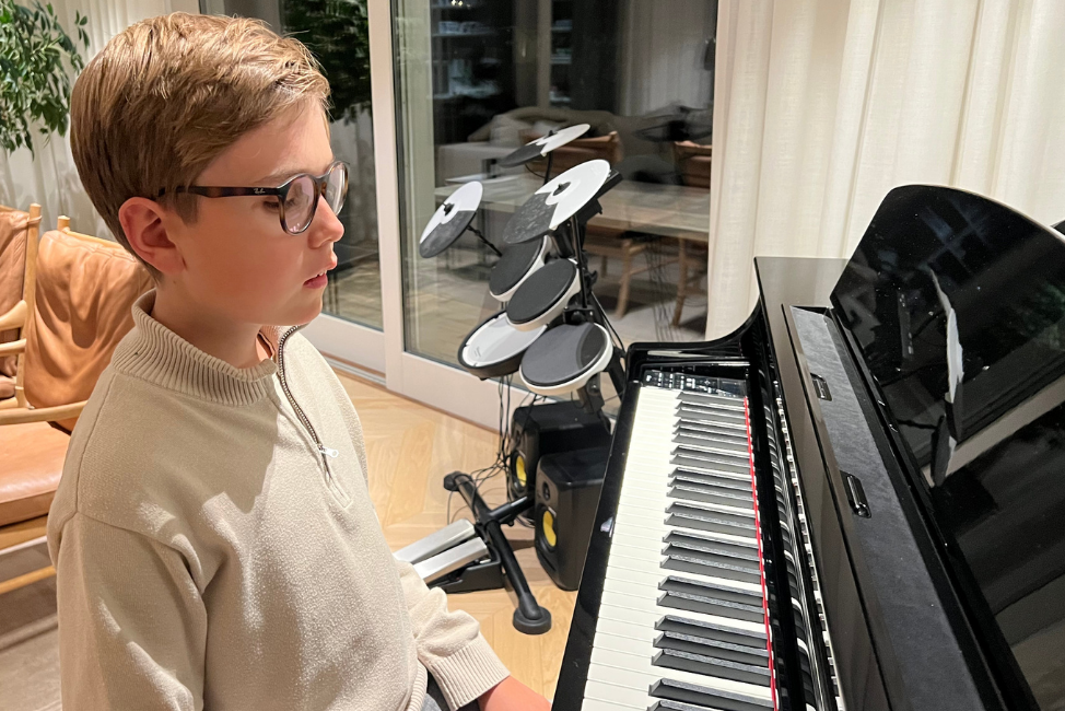 En ung blind, vit kille i beige tröja sitter vid ett svart piano i ett vardagsrum