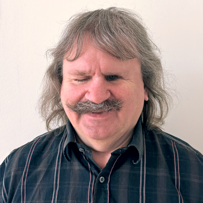 Porträtt av Håkan Thomson. Håkan är en man i 60-årsåldern med axellångt grått hår med lugg och en buskig mustasch. Han har en synlig synskada och ler.