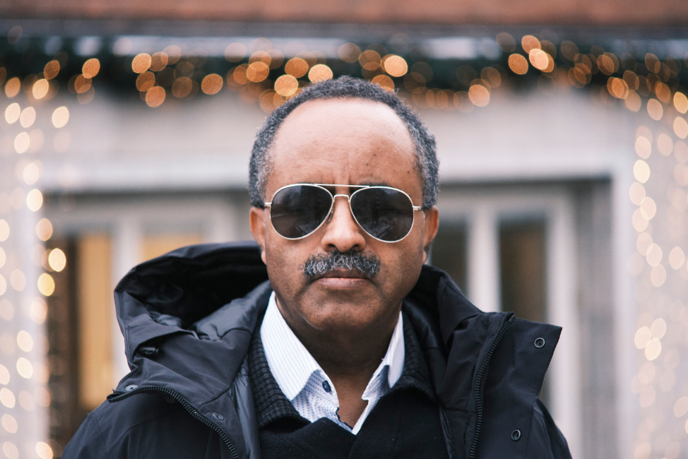 Tesfaye Deriba, En svart man, 65 år, med svarta pilotglasögon. Han har mustasch och snaggat hår, gråsprängt. Han bär en vit småprickig skjorta och över det en mörk tröja och en öppen svart jacka. Foto av Marcus Morey-Halldin