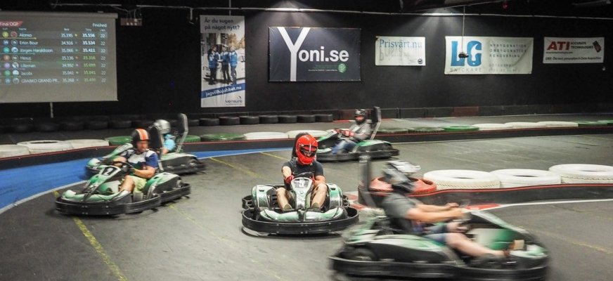Alt-text: Inomhusbild från en gokartbana på Gokartcentralen i Göteborg. Fem personer kör gokarts genom en kurva, alla med hjälmar och säkerhetsutrustning. Föraren i mitten bär en röd hjälm och ser ut att leda. Rörelseoskärpa syns på föraren i förgrunden som kör snabbt. I bakgrunden finns reklamskyltar på väggen och en digital resultattavla som visar förare och varvtider.