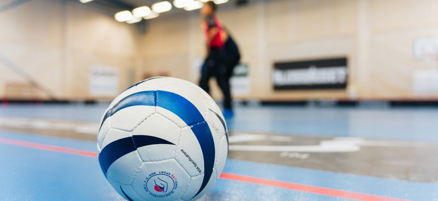 En fotboll i närbild. Den ligger på golvet på en 5-a-side-plan och i bakgrunden syns en spelare.