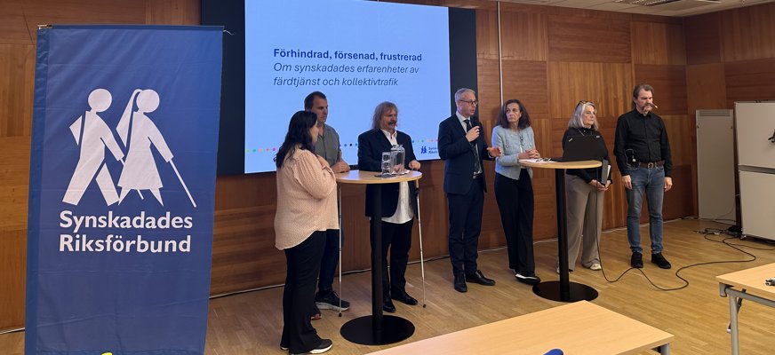 Sju personer står i en sal framför en stor skärm med texten "Förhindrad, försenad, frustrerad - Om synskadades erfarenheter av färdtjänst och kollektivtrafik".