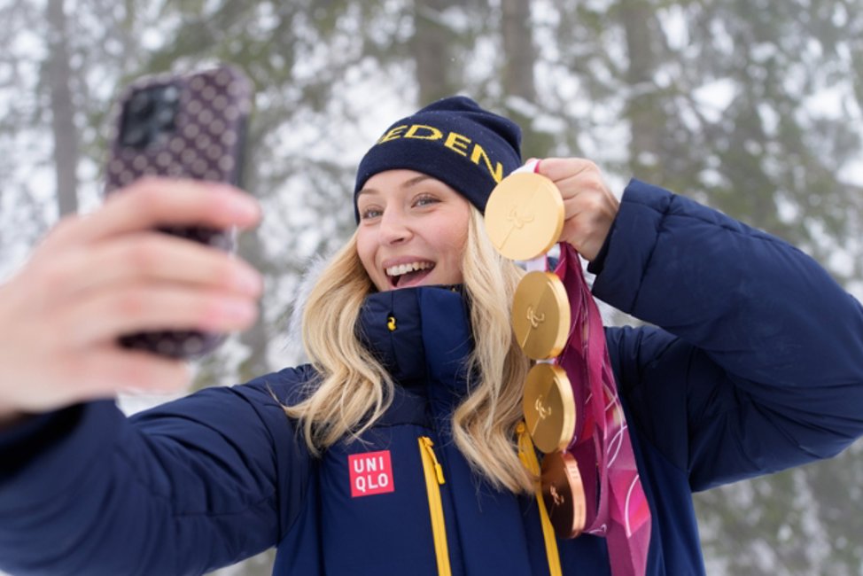En blond vit kvinna i 25-årsåldern tar en selfie med sin telefon. Hon bär en mörklblå mössa och en mörklblå jacka och visar upp fyra medaljer för kameran, 3 guld och en i brons. 