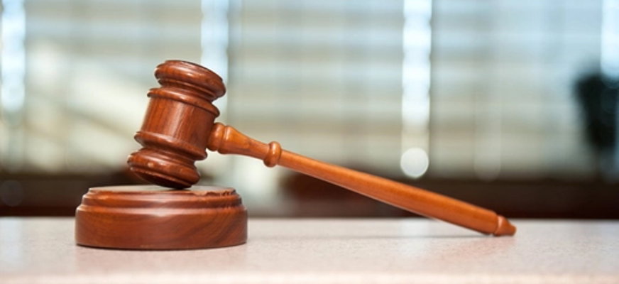 Bilden visar en mörkbrun ordförandeklubba i trä som ligger ovanpå en rund träplatta i samma färg. I bakgrunden ser man diffust några fönster med skira gardiner framför fönstren.