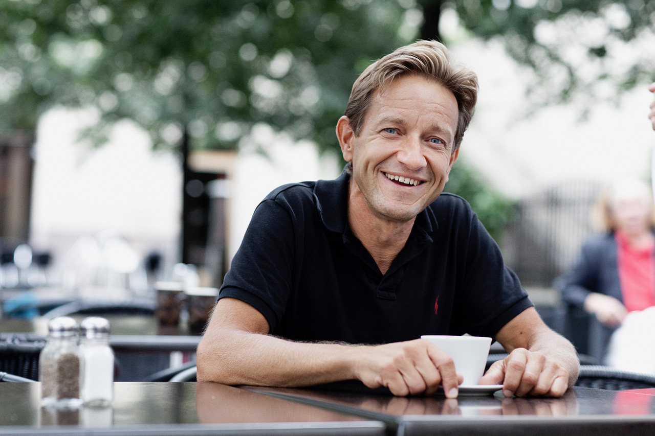 Anders Kvanta ler brett och har på sig en kortärmad, mörk pikétröja. Han sitter utomhus med en kopp kaffe vid ett kafébord.