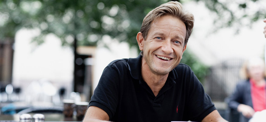 Anders Kvanta ler brett och har på sig en kortärmad, mörk pikétröja. Han sitter utomhus med en kopp kaffe vid ett kafébord.