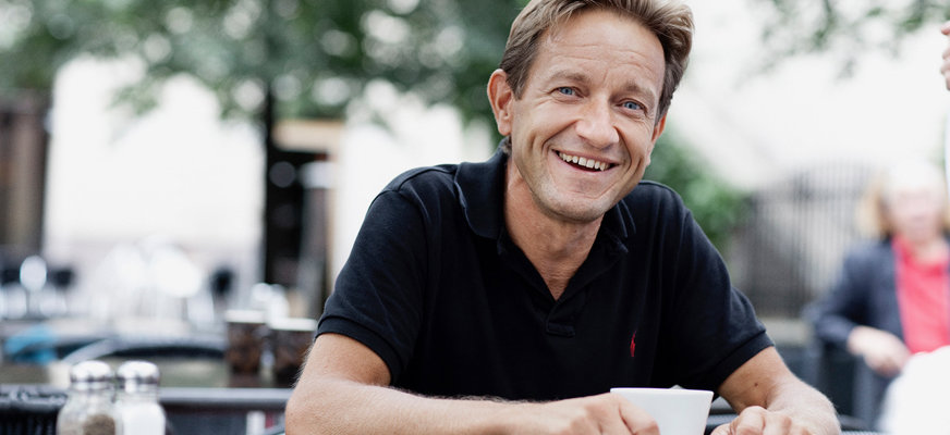 Anders Kvanta ler brett och har på sig en kortärmad, mörk pikétröja. Han sitter utomhus med en kopp kaffe vid ett kafébord.