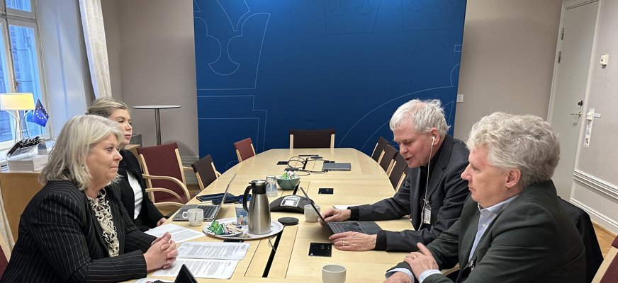 Camilla Waltersson Grönvall och politiskt sakkunnig Cassandra Lundgren sitter bredvid varandra vid ett mötesbord, iklädda mörka kavajer. Mitt emot sitter SRF:s ordförande Niklas Mattsson och sakkunnige Lennart Karlsson, de har både grått hår och mörka kavajer. De fyra ser allvarliga ut och samtalar med varandra, på borde ligger muggar, papper och datorer.