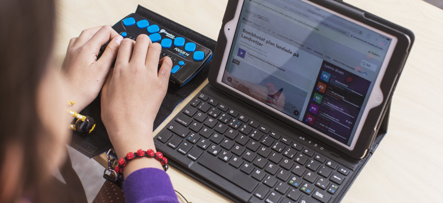 Händer som skriver på en punktskriftsdisplay placerad bredvid en datorskärm.