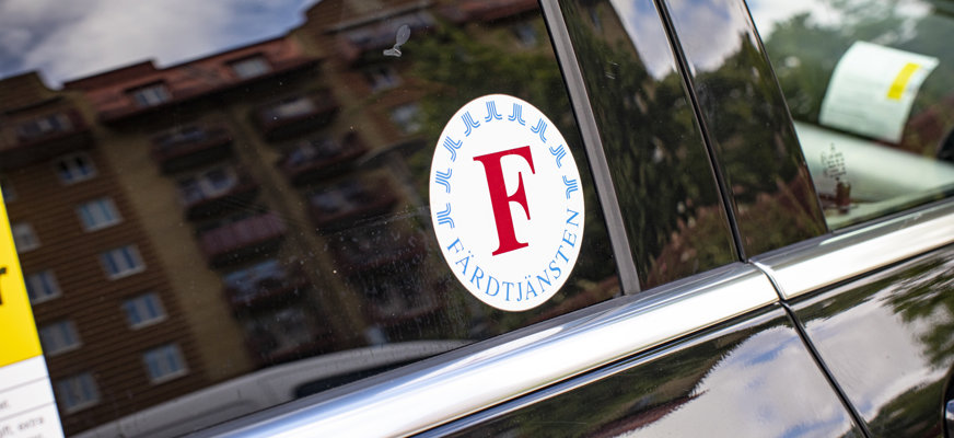 Närbild på svart färdtjänstbil. På rutan sitter ett klistermärke med färdtjänstens logotyp.