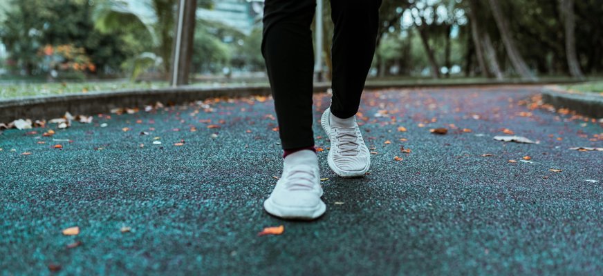 Två ben med svarta tights och vita sneakers. Personen är ute på promenad. På den gråblå asfalten ligger några orangea höstlöv utspridda.