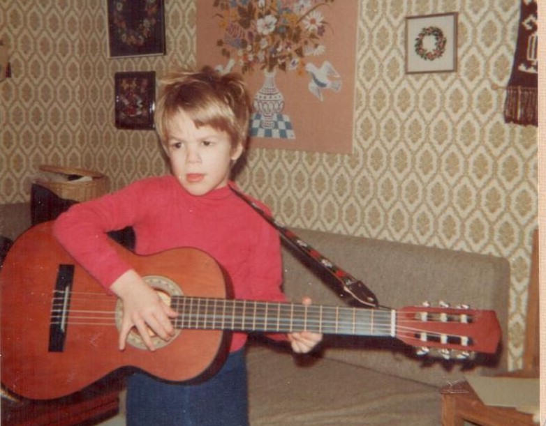 Fredrik som barn, en blond och brunögd pojke i femårsåldern, spelar gitarr. Hans kläder och rummets är inredning är i typisk sjuttiotalstil.