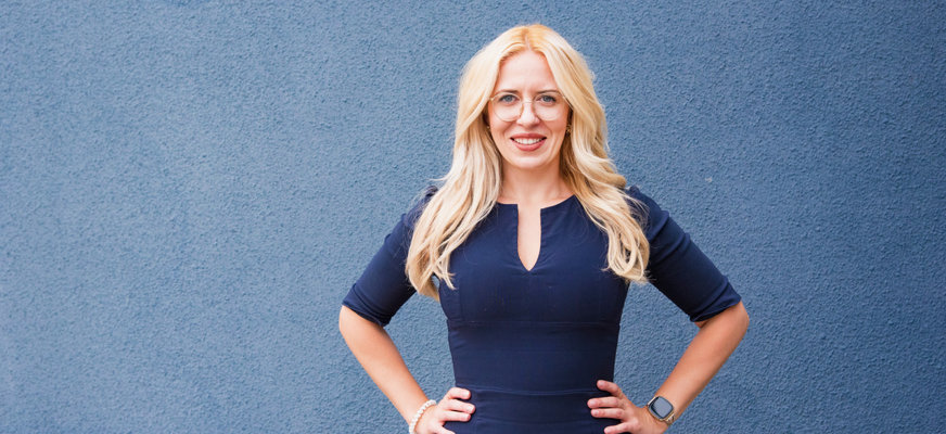 Andreea Badache har blont, långt hår och glasögon med tunna bågar. Hon ler stort, har på sig en blå klänning och står upp med händerna på höfterna.