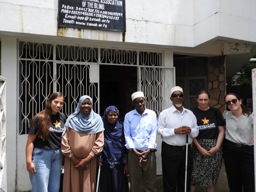 Sju personer står uppradade utanför Zanzibar National Association of the blind (ZANAB). Fyra av dem är från organisationen och står med vita käppar. Två av dem är från My Right och en från SRF. 