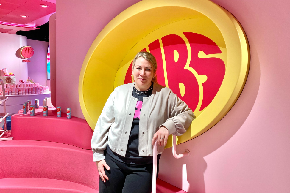 Joanna Mossberg, kommunikationschef BUBS. 35 årig kvinna med blont hår i tofs, beige kofta och svart t-shirt under. Står vid rosa vägg med stor logotype av ”BUBS”- rosa bokstäver i ljusgul oval. Foto: Aziza Dhaouadi