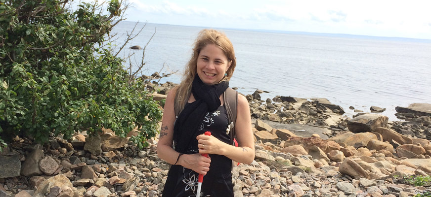 Ulrika Wiland står med vit käpp på en stenstrand med havet i bakgrunden. Det är sommar och hon har svart sommarklänning och svart sjal.
