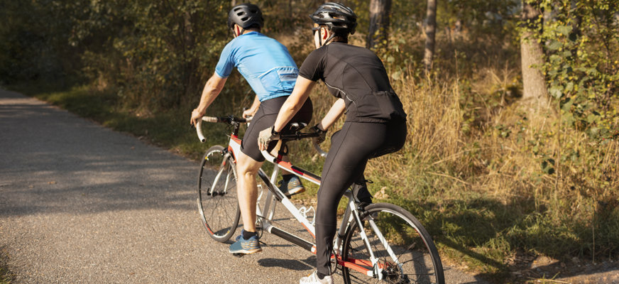 Två personer cyklar på en tandemcykel längs en grusväg i skogen. Solen skiner och vägen kantas av träd och grönska.