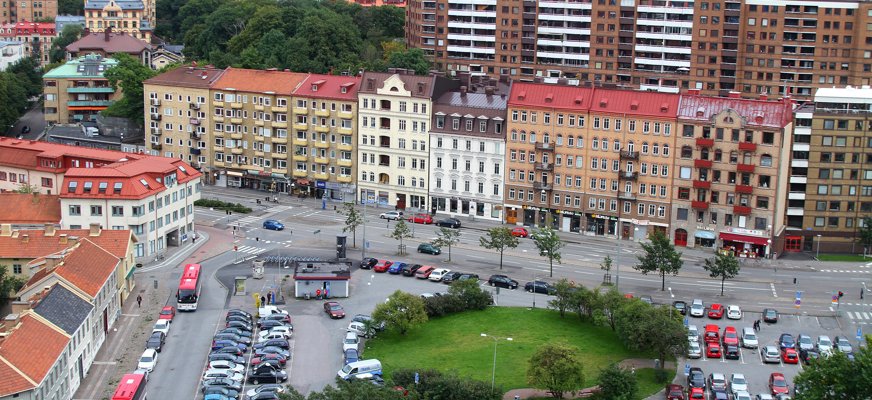 Flygfoto som visar Skanstorget i Göteborg med en rund gräsyta, parkerade bilar, bussar och flerbostadshus längs torget.