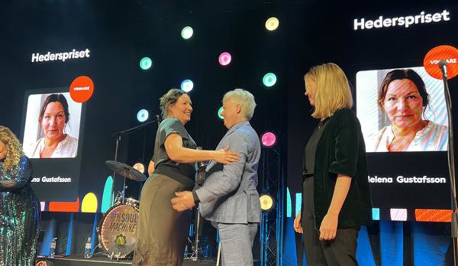 Helena Gustafsson mottar hederspriset av Niklas Mattsson på scenen. De kramas. Bakom dem syns Helenas ansikte på storbildsskärmar.