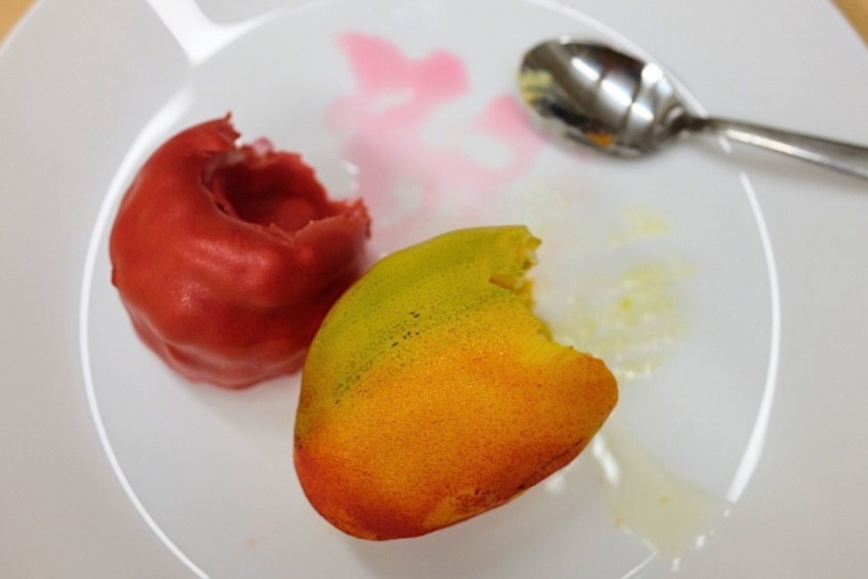 På en vit tallrik ligger två bakelser som ser ut som en gul mango och ett rosa stort hallon