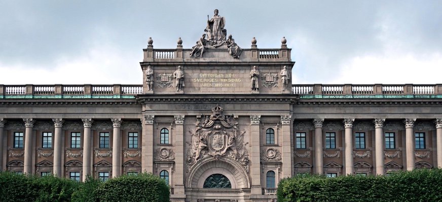 Riksdagshuset i Stockholm fotat framifrån. Porten är maffig med stora pelare omkring sig och framför huset grönskar det.