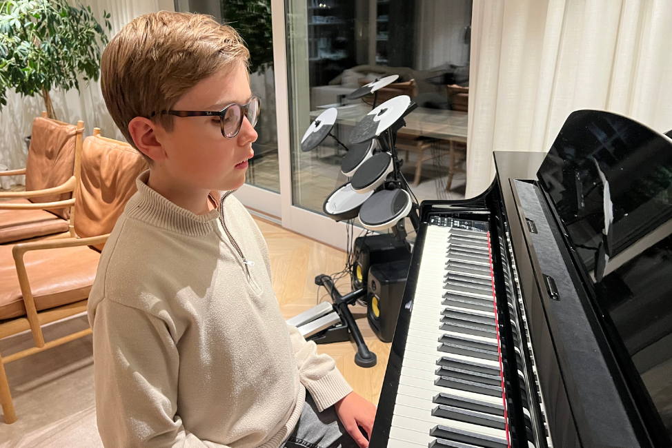 En ung blind, vit kille i beige tröja sitter vid ett svart piano i ett vardagsrum