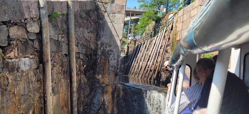 Båt i en sluss på Dalslands kanal med stenmurar, rinnande vatten och passagerare som tittar ut i solsken.