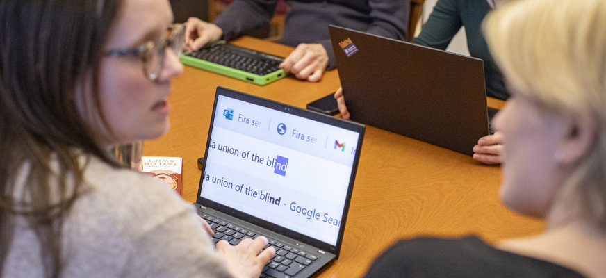 En grupp personer sitter i ett konferensrum. Två personer i förgrunden diskuterar något över en dataskärm med förstoring.