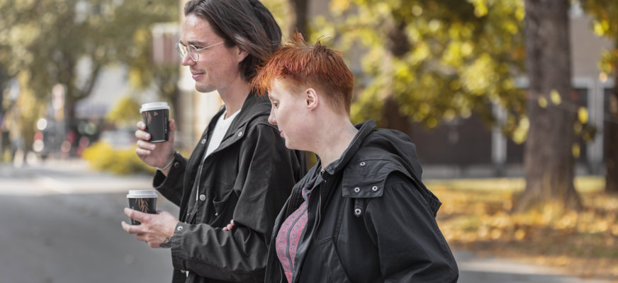 Kvinna med vit käpp ledsagas av person med två kaffemuggar i händerna