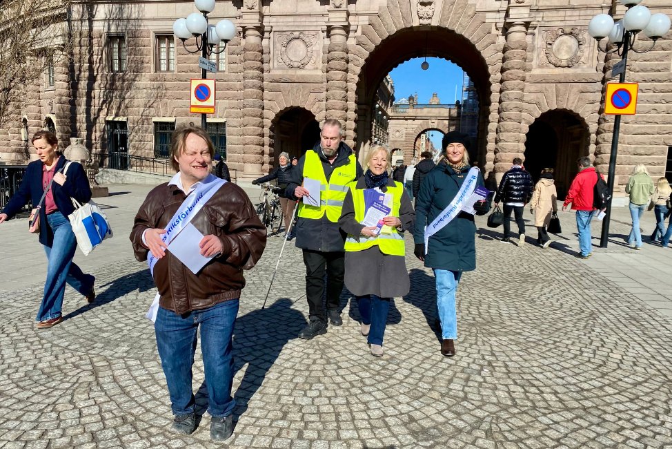 På Mynttorget vid riksdagshuset, går några personer med SRF ordensband i vitt och har broschyrer som de delar ut till förbipasserande.