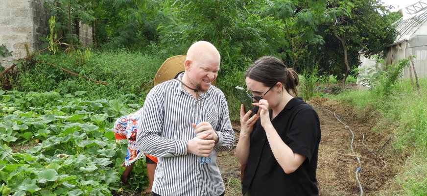 Två personer står i ett grönsaksland och kollar på en telefon tillsammans. Den ena personen är svensk och kollar på sin mobilskärm med förstoring. Den andra personen är Tanzanian och har albinism.