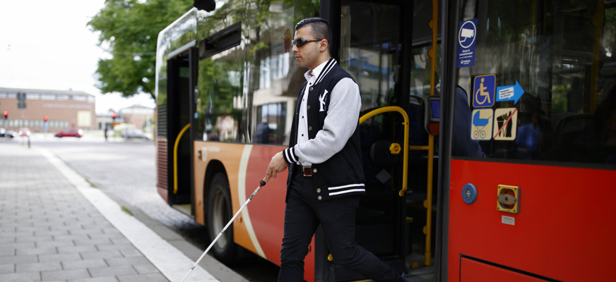 En man med svart hår, vit och svart jacka samt solglasögon och vit käpp kliver ur en buss på en stadsgata. I bakgrunden syns ett träd och en låg byggnad.