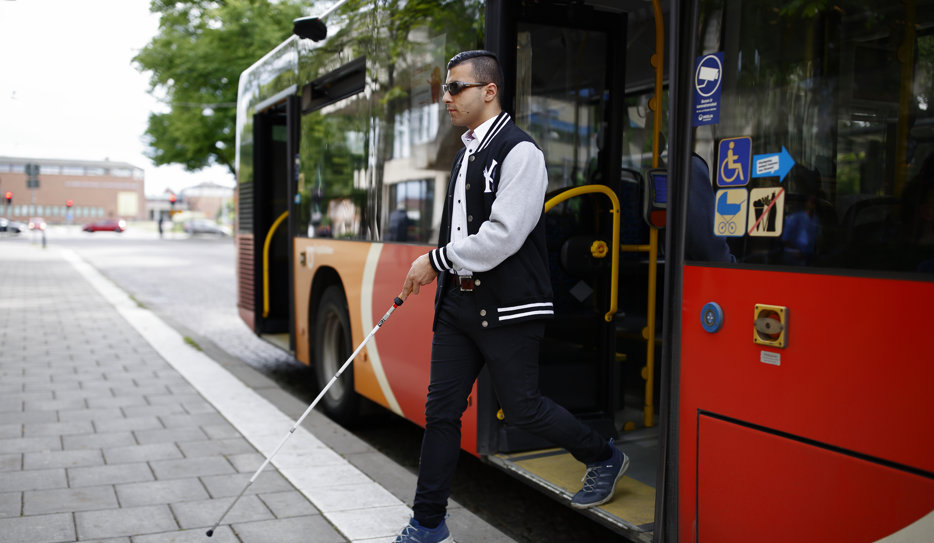 En man med svart hår, vit och svart jacka samt solglasögon och vit käpp kliver ur en buss på en stadsgata. I bakgrunden syns ett träd och en låg byggnad.