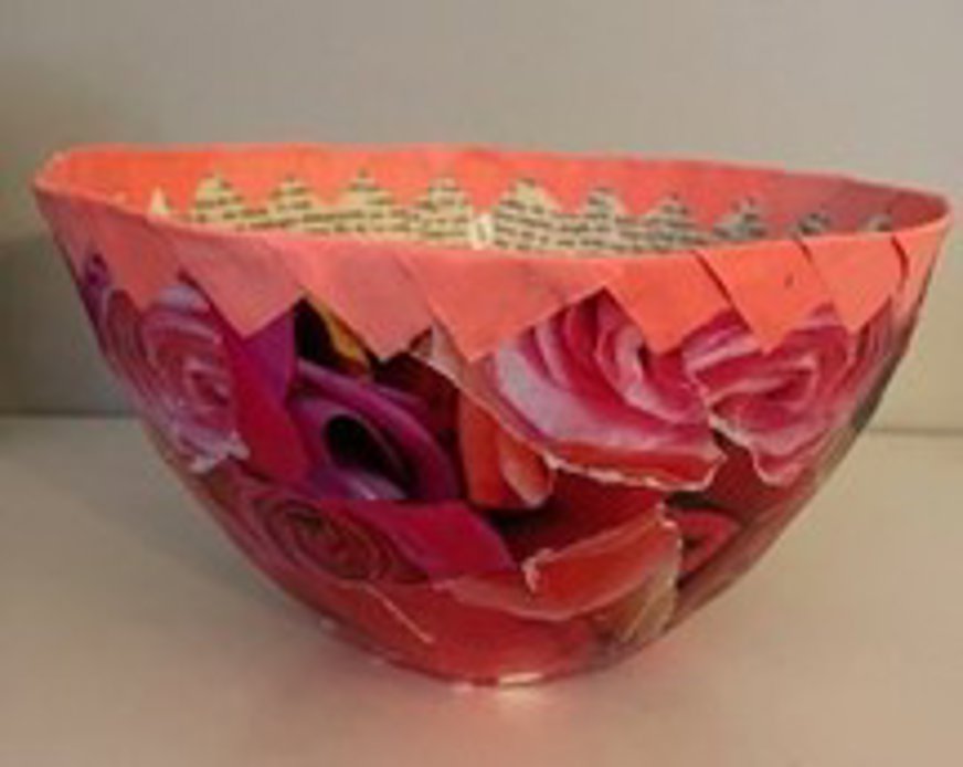 Skålen är gjord i collage-teknik med färgstarka bilder av rosor och andra blommor som täcker hela ytan. Den har rund form och är uppbyggd av papper eller liknande lätt material. Överkanten pryds av en rosa pappersremsa med sicksackmönster, där tryckt text syns på insidan. Färgerna är livliga med dominans av rött, rosa och orange, vilket ger ett uttrycksfullt och dekorativt intryck. Skålen står mot en neutral bakgrund som framhäver dess detaljer.